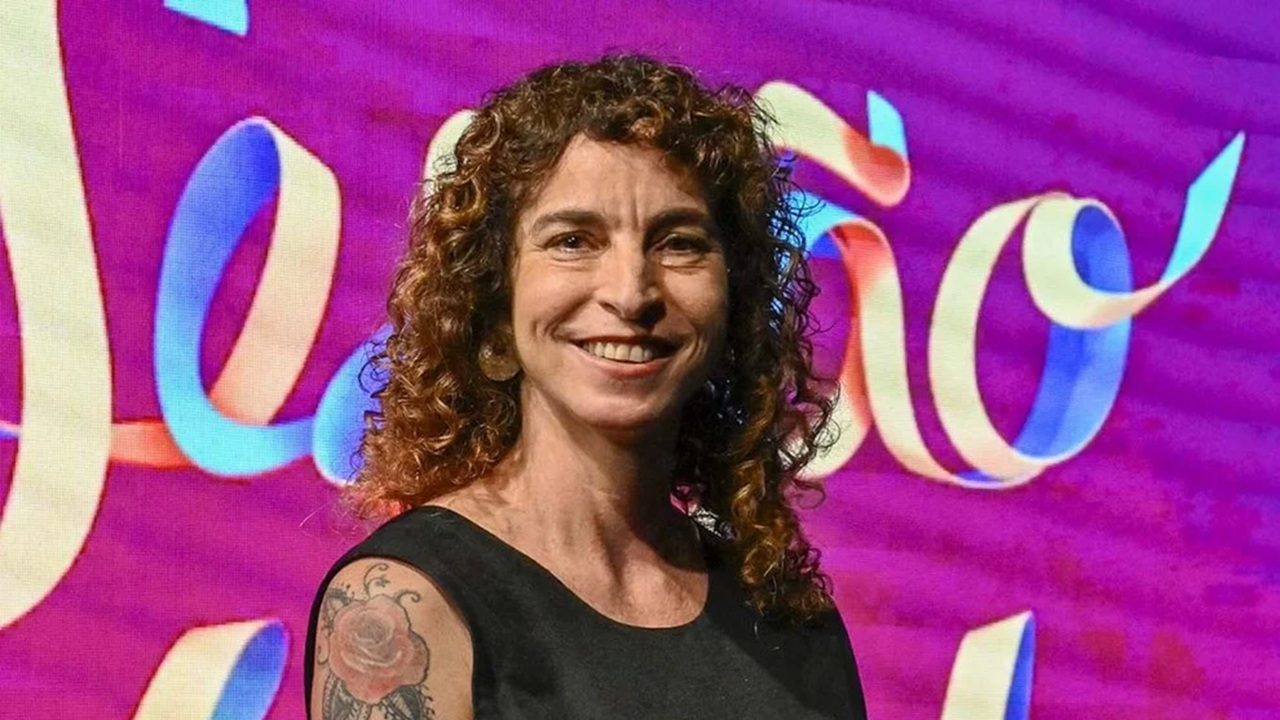 Mulher de cabelos cacheados castanhos e sorriso aberto posa em frente a um painel colorido com tons de roxo, rosa e letras em azul e dourado. Ela usa vestido preto sem mangas e exibe uma tatuagem de rosa no ombro esquerdo.