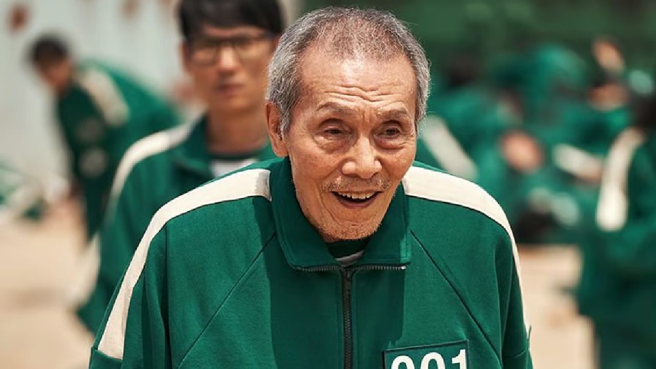 Cena de série Round 6 mostra um homem idoso vestindo uniforme verde numerado “001”, em um ambiente externo com outras pessoas ao fundo usando roupas semelhantes