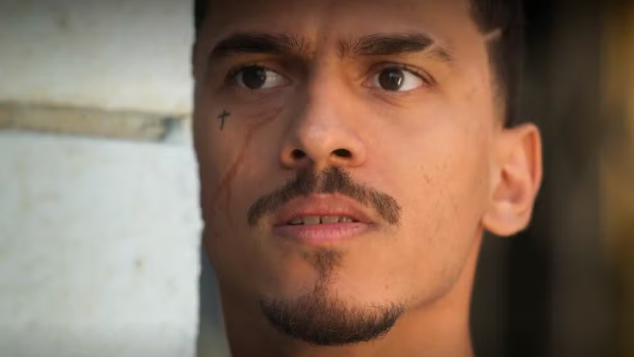 A imagem mostra um homem de expressão séria, encostado em uma parede clara. Ele tem um pequeno bigode e cavanhaque, além de uma tatuagem em forma de cruz próxima ao olho e uma cicatriz que desce pelo lado esquerdo do rosto. A iluminação destaca seu olhar atento e concentrado, sugerindo tensão ou expectativa na cena