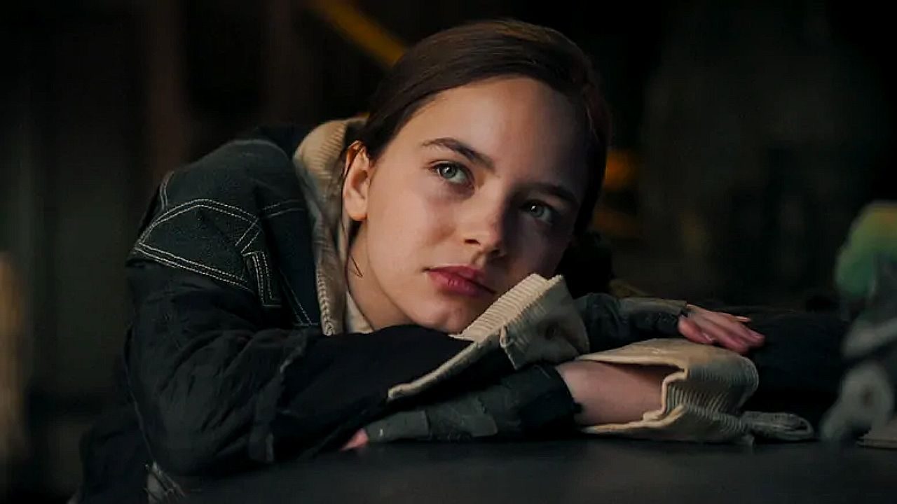 Uma jovem está apoiada sobre uma mesa, com a cabeça descansando sobre os braços cruzados. Ela usa um casaco escuro com capuz e uma blusa clara por baixo. Seu olhar está voltado para o lado, com expressão pensativa ou contemplativa. A iluminação é suave e o ambiente ao fundo é escuro e desfocado, sugerindo um espaço interno e silencioso, possivelmente um quarto ou sala pouco iluminada.