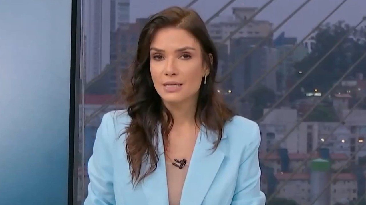 Sabina Simonato de blazer azul-claro apresenta o Bom Dia São Paulo diante de janela com vista da cidade