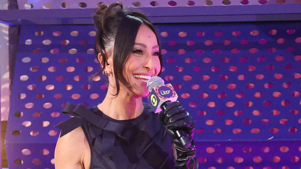 Sabrina Sato sorrindo e segurando microfone com logo da Globo no Festival LED diante de painel roxo iluminado
