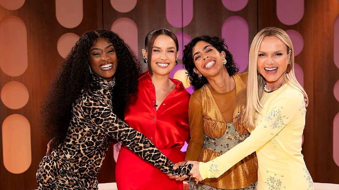 Apresentadoras sorrindo de mãos dadas no palco de programa de TV com cenário colorido ao fundo