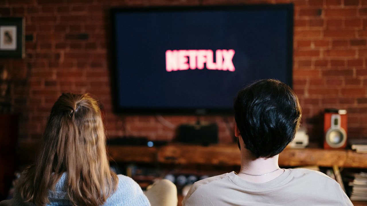 Casal sentado de costas assistindo à tela de televisão com logotipo da Netflix exibido