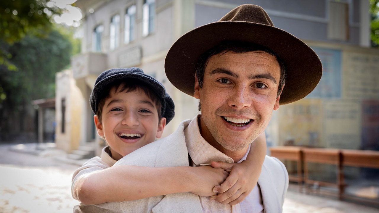 Samir (Davi Malizia) abraçado nas costas de Candinho (Sergio Guizé) sorrindo em cena da novela Êta Mundo Melhor!