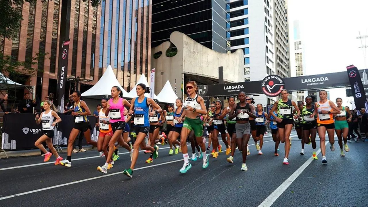 Grupo de corredoras participa de uma prova de rua em ambiente urbano com prédios altos ao fundo, passando pelo pórtico de largada onde se lê “Largada” e “São Silvestre”, com público e organizadores observando nas laterais da avenida