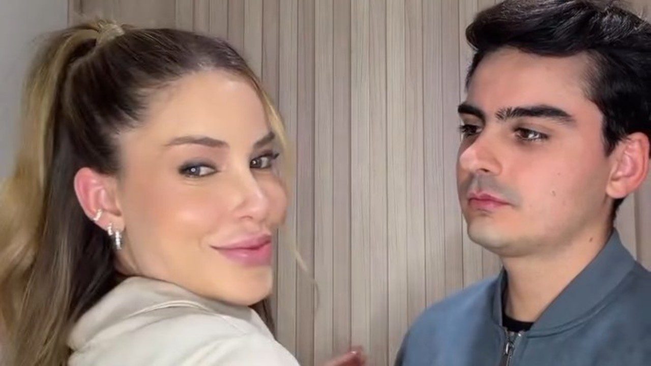 Saory Cardoso sorri para a câmera de blazer claro ao lado de Dudu Camargo de jaqueta cinza diante de parede ripada