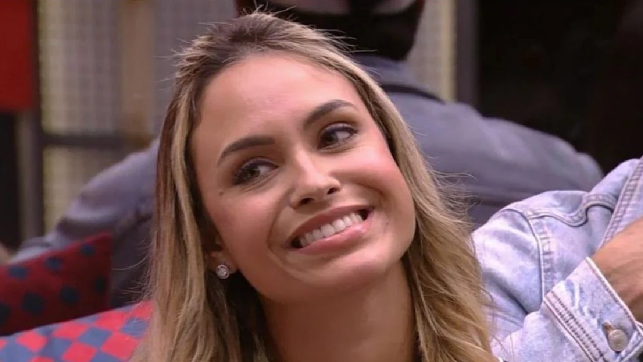 A imagem mostra uma mulher sorrindo, com cabelos loiros e lisos, usando brincos pequenos e redondos. Ela está em um ambiente interno, com pessoas e móveis visíveis ao fundo, incluindo uma almofada com estampa vermelha e azul