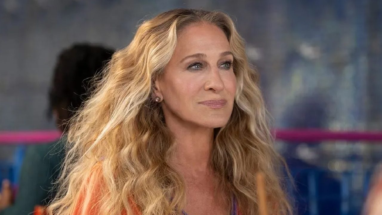 Mulher de cabelos longos e ondulados, loiros, com expressão serena, olhando para a frente, em ambiente externo desfocado, vestindo roupa em tom quente e maquiagem discreta