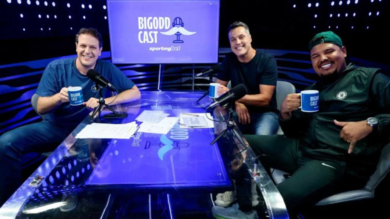 Três homens sorrindo seguram canecas do Bigodd Cast em mesa de estúdio com microfones e telão do podcast ao fundo
