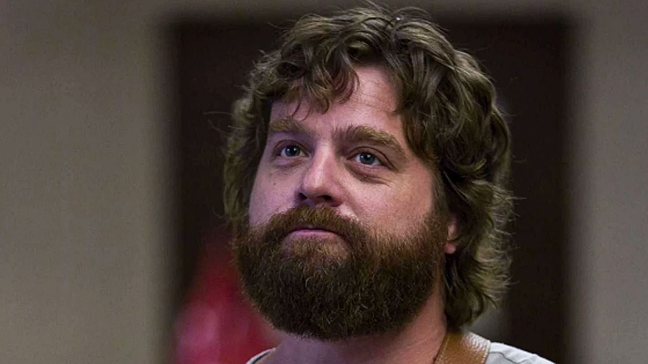 Homem de barba longa e cabelo cacheado observa alguém em cena de série de comédia dramática
