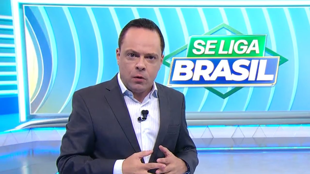 Thiago Gardinali, apresentador do jornal Se Liga, Brasil, gesticula em frente ao telão com logo do programa no estúdio do SBT.