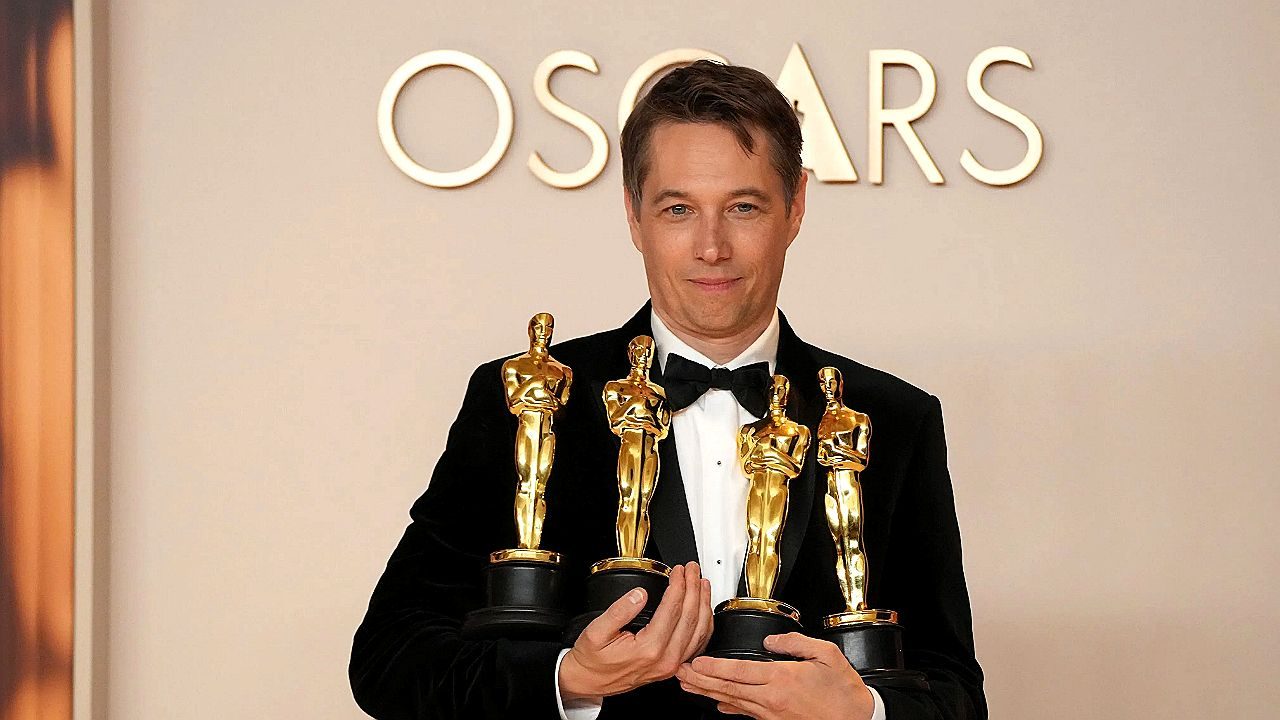 Homem de terno segura quatro estatuetas do Oscar durante premiação com painel escrito Oscars ao fundo