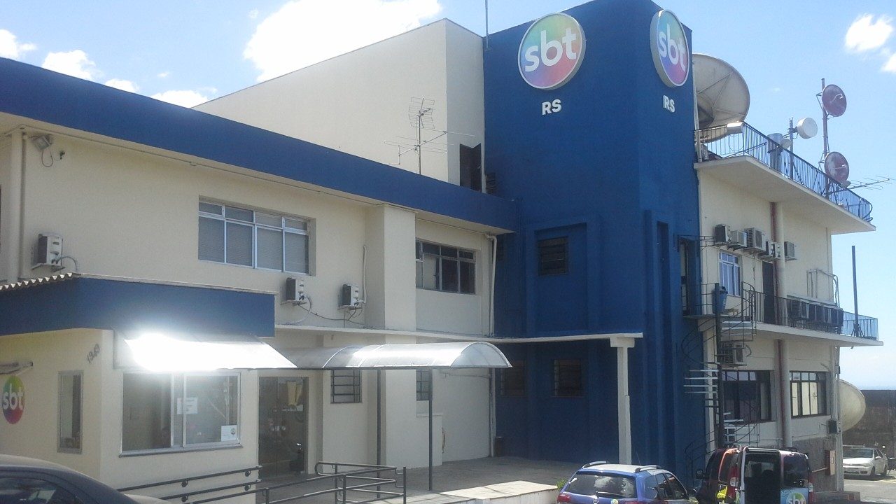 Fachada da sede do SBT RS com logotipos da emissora em prédio de dois andares e antenas de transmissão no topo