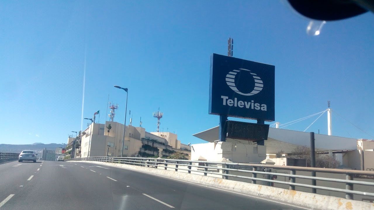 Placa com logotipo da Televisa ao lado de rodovia elevada, com sede da emissora e torres de transmissão ao fundo