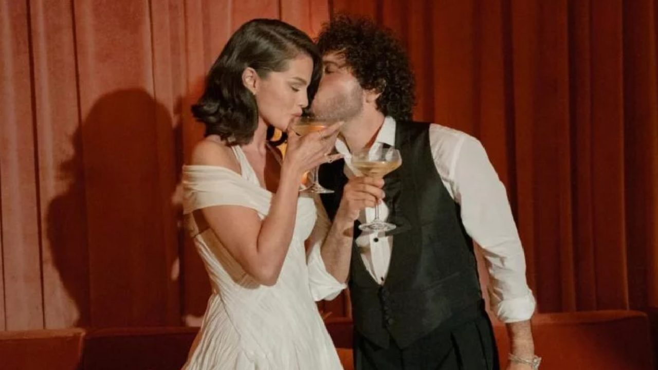 A imagem mostra um casal brindando com taças de champanhe em um ambiente elegante, com cortinas vermelhas ao fundo. A mulher usa um vestido branco de ombro caído e cabelo curto penteado em ondas, enquanto o homem, de colete preto e camisa branca