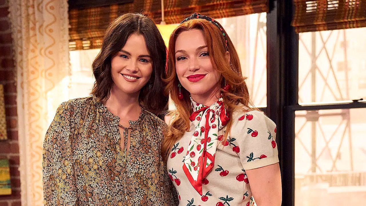 Selena Gomez e outra atriz posam sorrindo juntas em cena de série ambientada em apartamento com decoração retrô