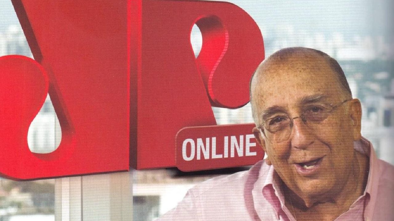 Homem mais velho, sorrindo, usando camisa rosa e óculos, posando diante de uma grande logomarca vermelha com as letras estilizadas “JP” e o termo “ONLINE”. Ao fundo, vê-se uma paisagem urbana desfocada através de janelas amplas