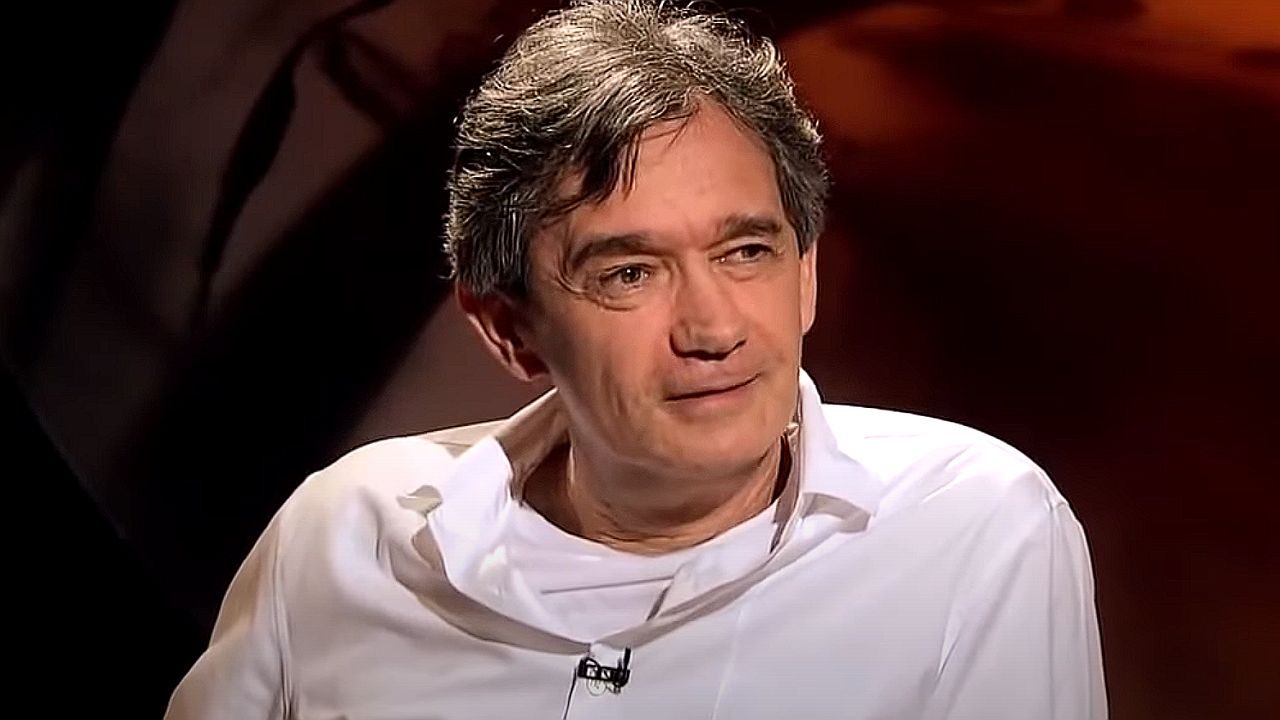 Homem de camisa branca participa de entrevista em estúdio com iluminação baixa e fundo avermelhado