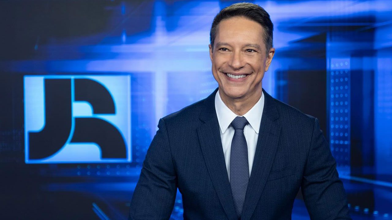 Sérgio Aguiar sorrindo no estúdio do Jornal da Record com logotipo do telejornal ao fundo