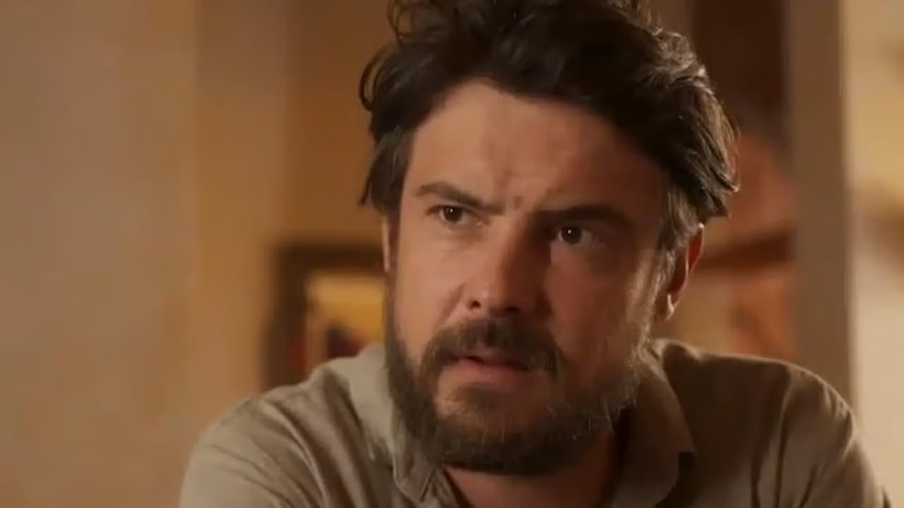 Homem de barba e camisa clara com expressão séria em ambiente interno iluminado, em cena de dramaturgia televisiva
