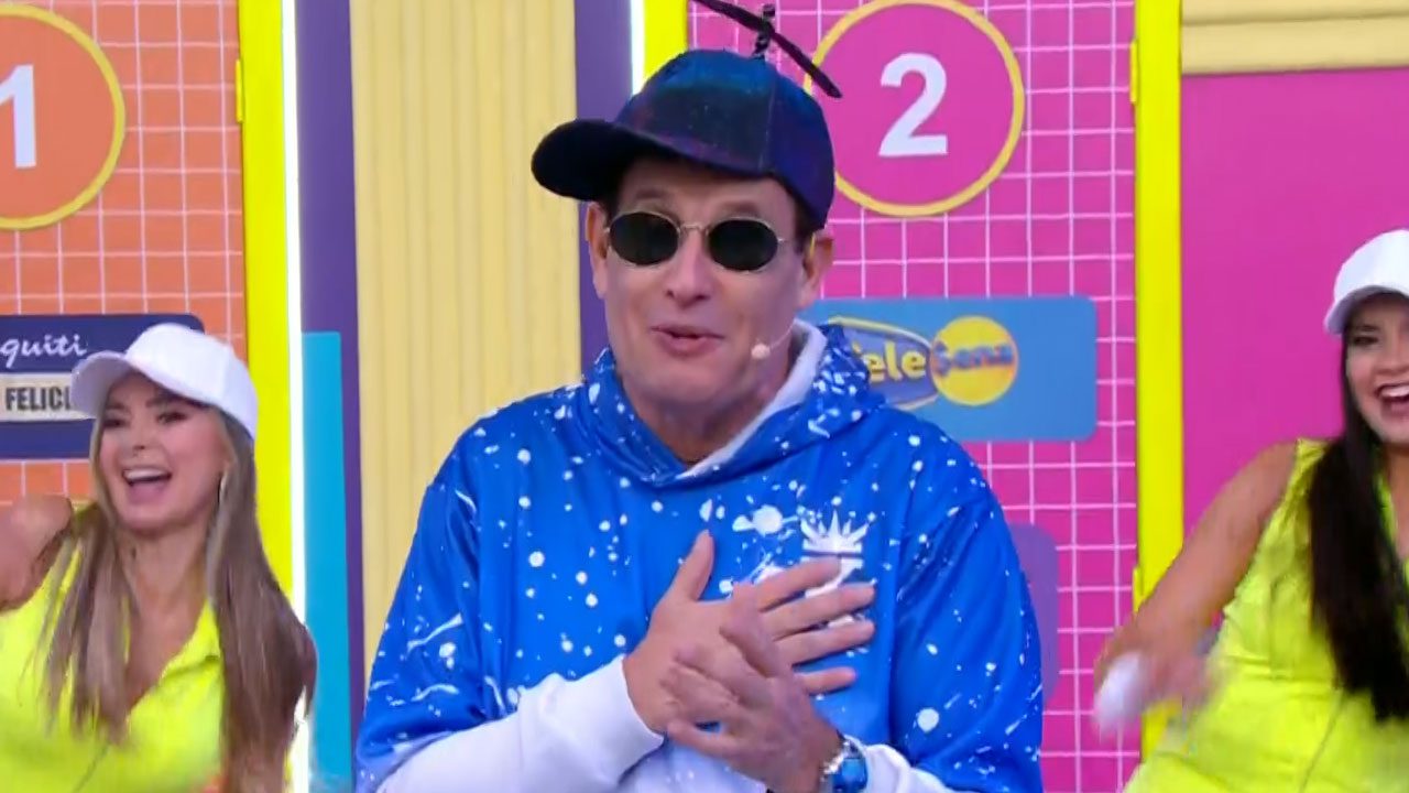 Sérgio Mallandro com boné de hélice óculos escuros e casaco azul apresenta programa de auditório com dançarinas ao fundo