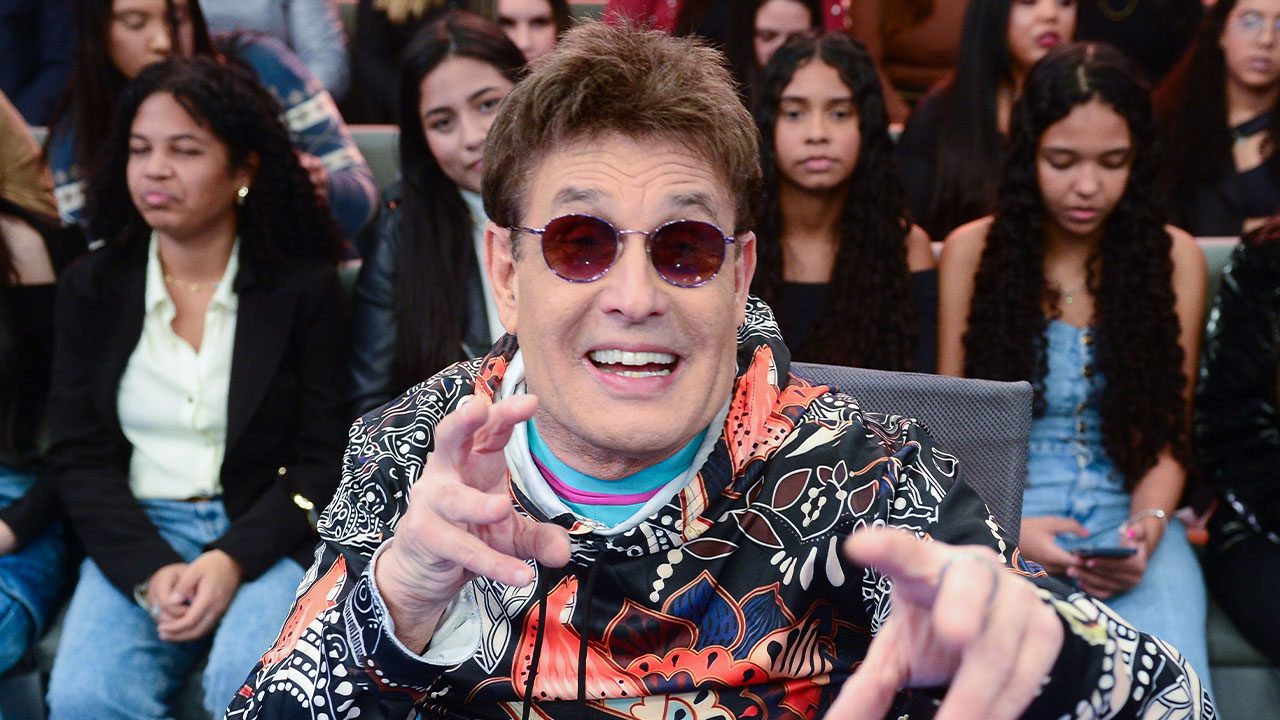 Sérgio Mallandro com óculos escuros e blusa estampada colorida gesticula diante de plateia no Programa do Ratinho