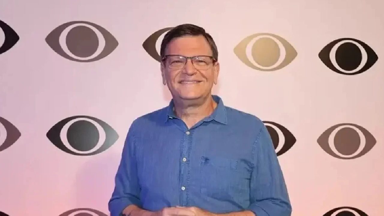 Homem sorri para a câmera diante de um painel com o logotipo repetido de uma emissora de televisão, vestindo camisa azul e óculos, em ambiente de evento ou estúdio