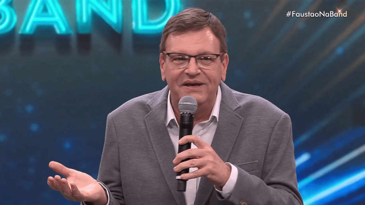 Homem de meia-idade falando em um microfone em um estúdio de TV. Ele veste blazer cinza e camisa clara, usa óculos e faz um gesto com a mão esquerda enquanto olha para a plateia ou câmera. O fundo é azul com luzes e elementos gráficos, e no canto superior direito aparece a hashtag “#FaustaoNaBand”.