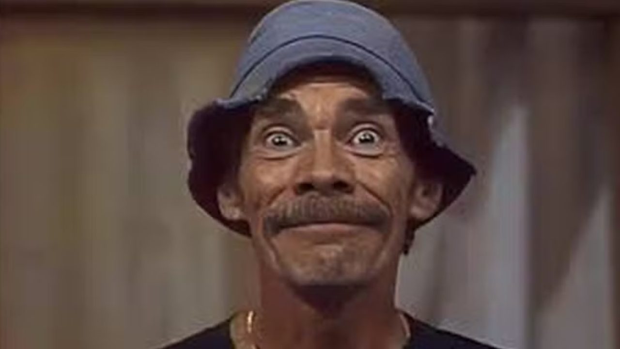 Homem de chapéu azul sorri com olhos arregalados em ambiente interno de cenário televisivo