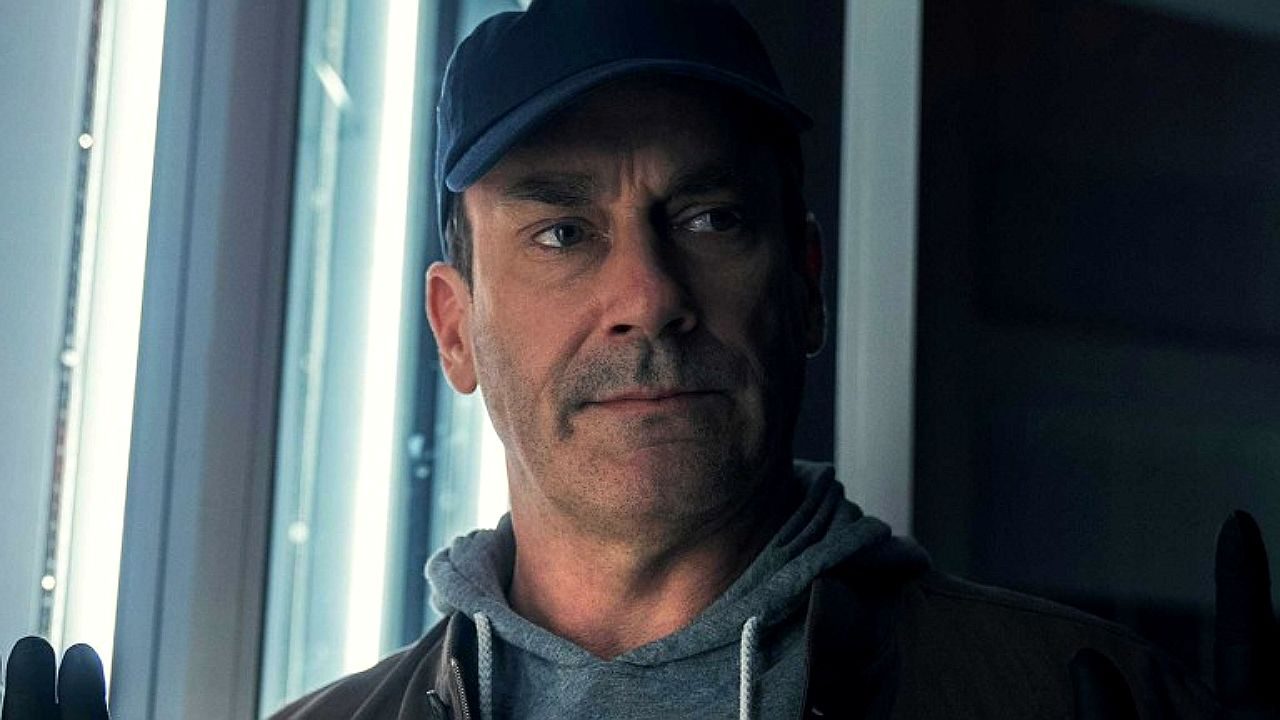 Homem de boné e moletom observa pela janela com expressão séria em cena de série dramática