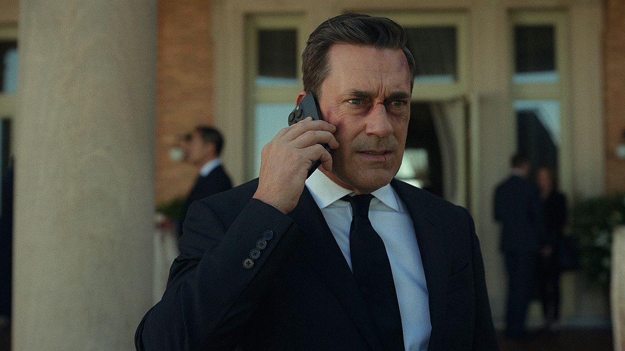 Homem de terno fala ao telefone com expressão séria em frente a prédio com pessoas ao fundo