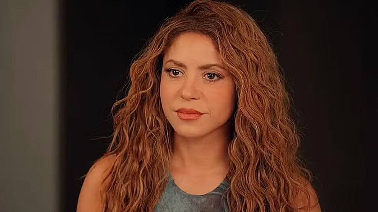 Shakira com cabelo cacheado e expressão séria em cena de série dramática durante conversa dentro de casa