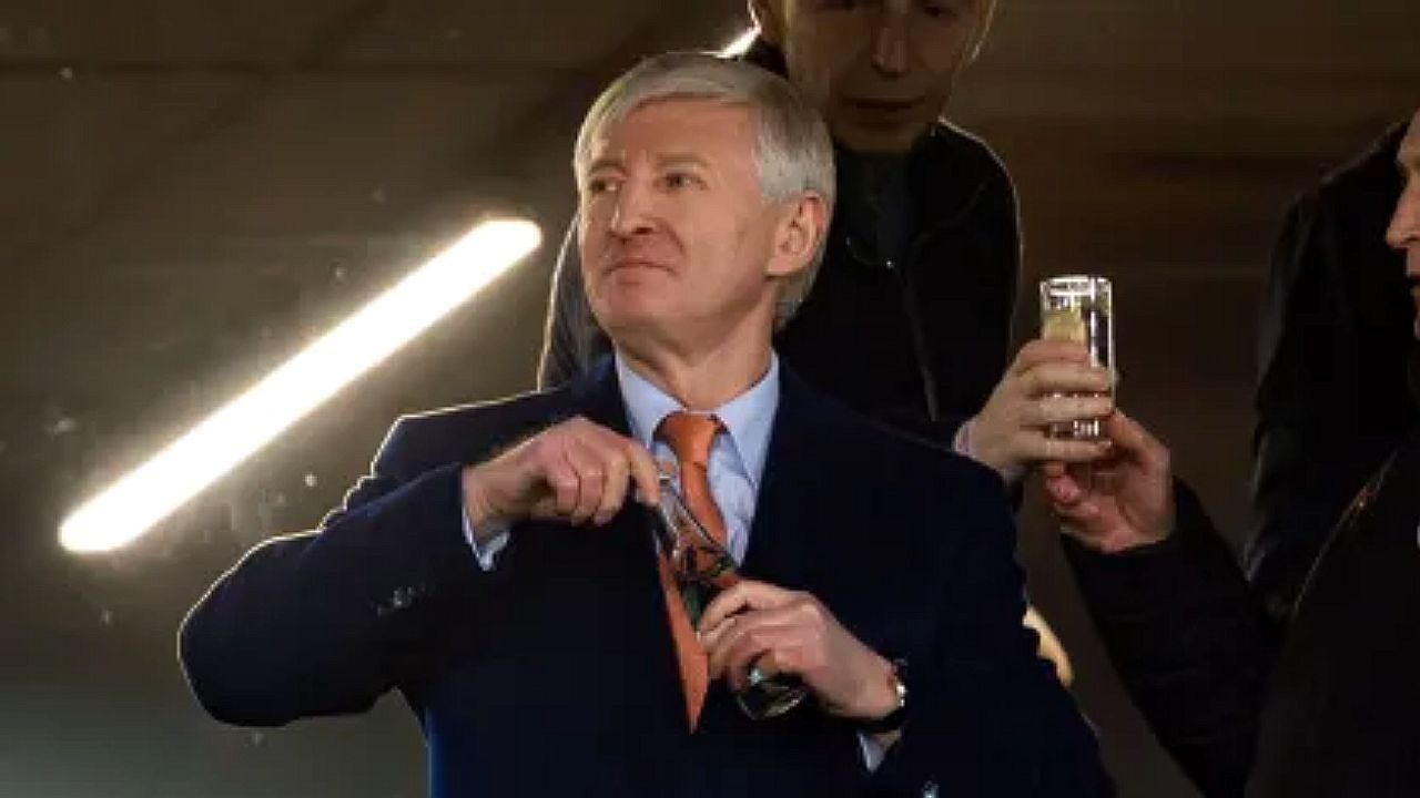 Homem de terno e gravata abre garrafa enquanto outro segura copo em cena com iluminação de estúdio ao fundo