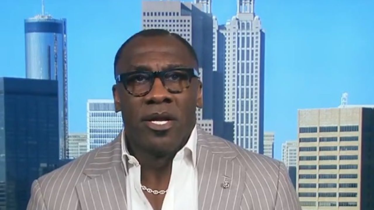 Shannon Sharpe com paletó listrado e óculos falando em participação na ESPN com prédios ao fundo
