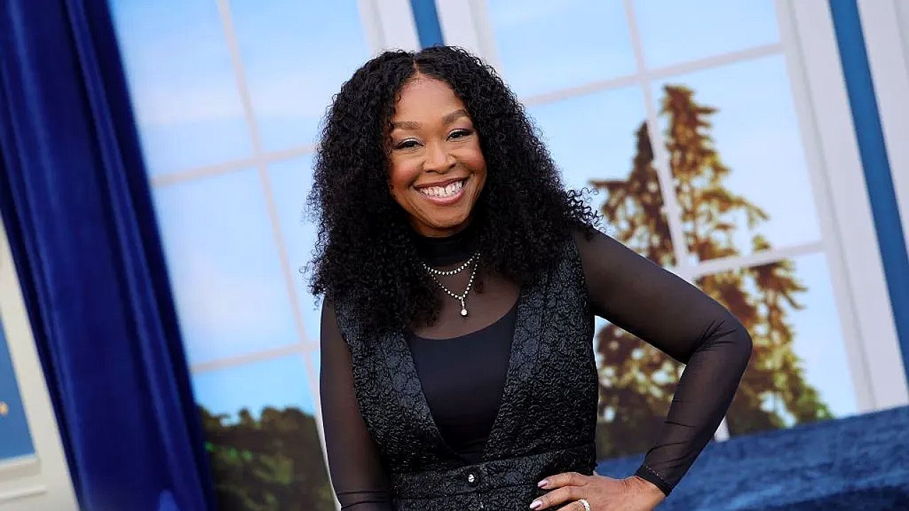 Uma mulher com cabelos longos e cacheados sorri enquanto está em um estúdio de televisão. Ela usa uma blusa preta de mangas transparentes com um colete escuro por cima, além de colares delicados. Está com uma das mãos apoiada na cintura, em uma postura confiante e descontraída. Ao fundo, há um cenário que imita grandes janelas com vista para árvores e céu, além de cortinas azuis, sugerindo um ambiente de programa de entretenimento ou talk show.