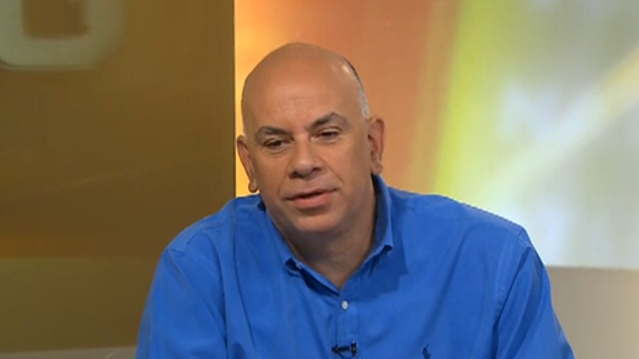 Homem de camisa azul participa de programa de TV, com fundo em tons de amarelo e laranja