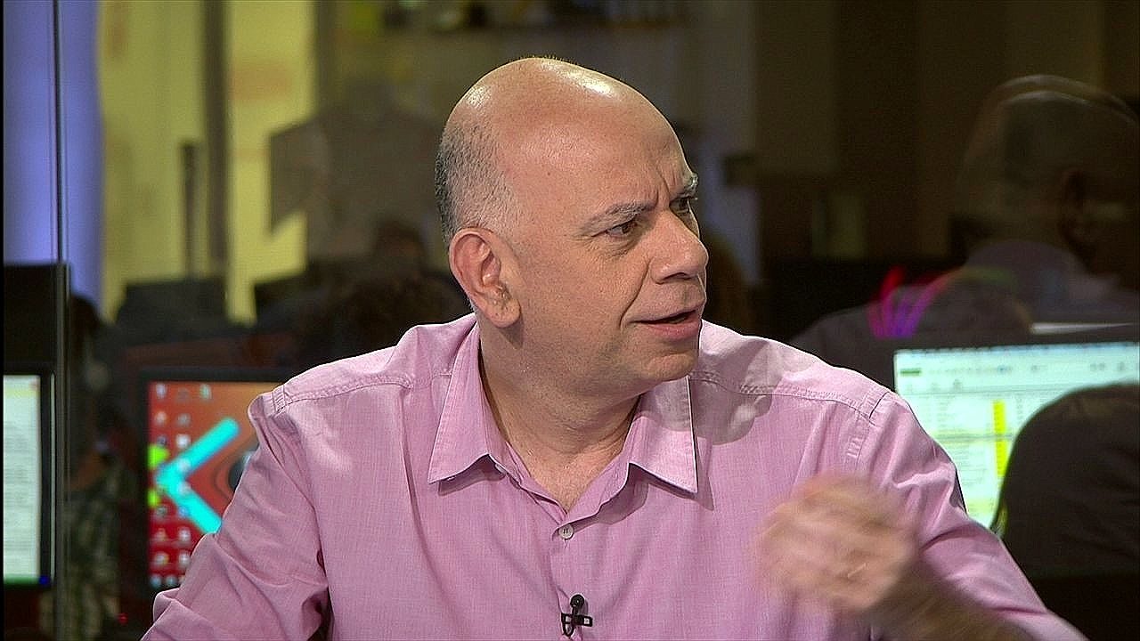 Homem careca veste camisa rosa e conversa em estúdio de televisão ou redação, com computadores e monitores ao fundo enquanto gesticula durante a fala.