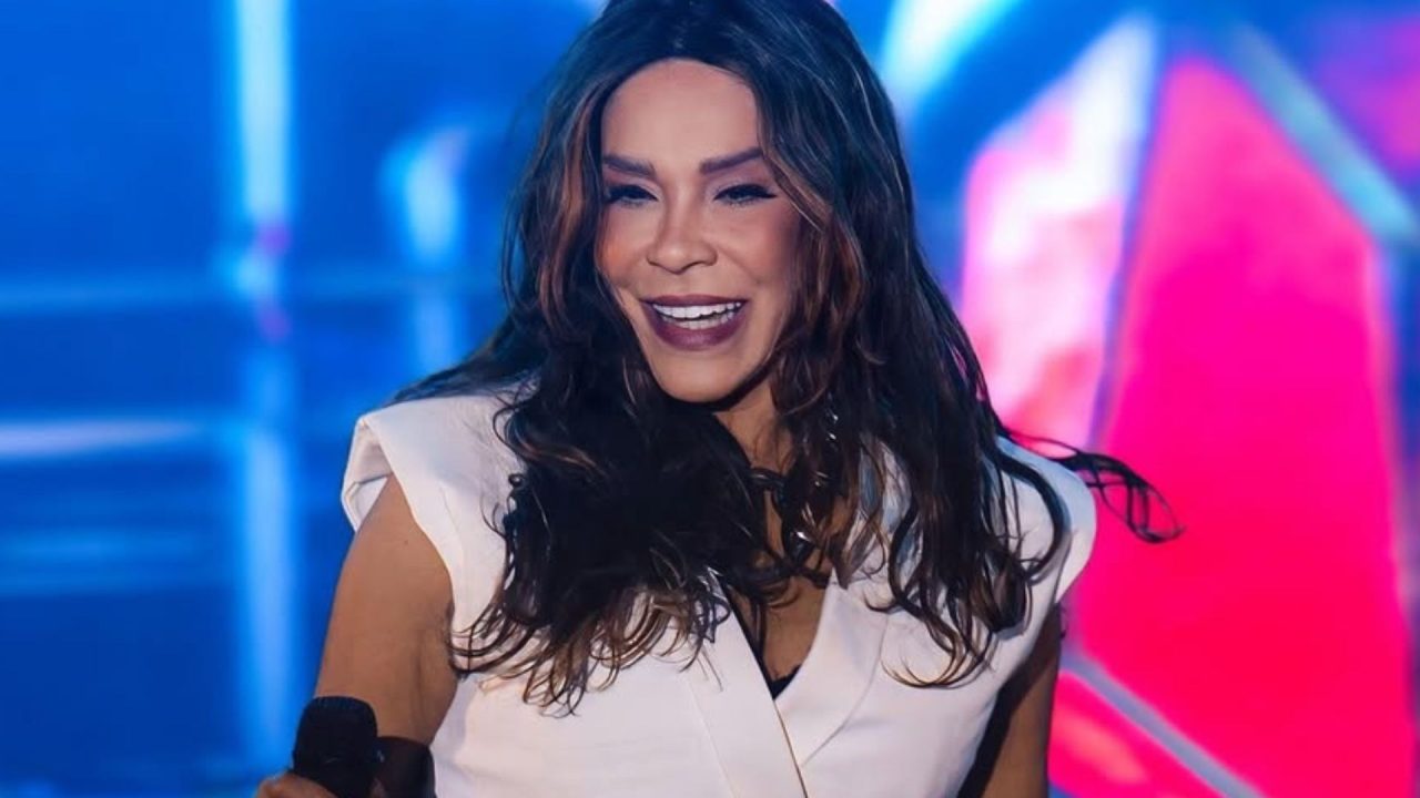 Cantora sorri enquanto segura microfone em apresentação ao vivo, com luzes coloridas e cenário vibrante ao fundo