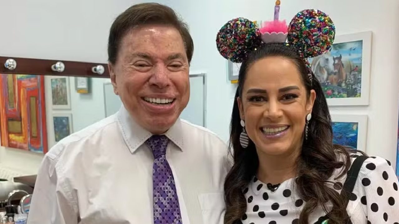 Homem de camisa social e gravata posa sorridente ao lado de mulher com tiara colorida em formato de orelhas e roupa de bolinhas, ambos em ambiente interno decorado com quadros coloridos