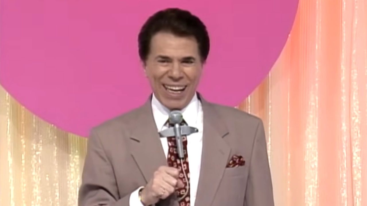 Silvio Santos sorrindo no palco do Topa Tudo Por Dinheiro com fundo rosa e cortinas brilhantes