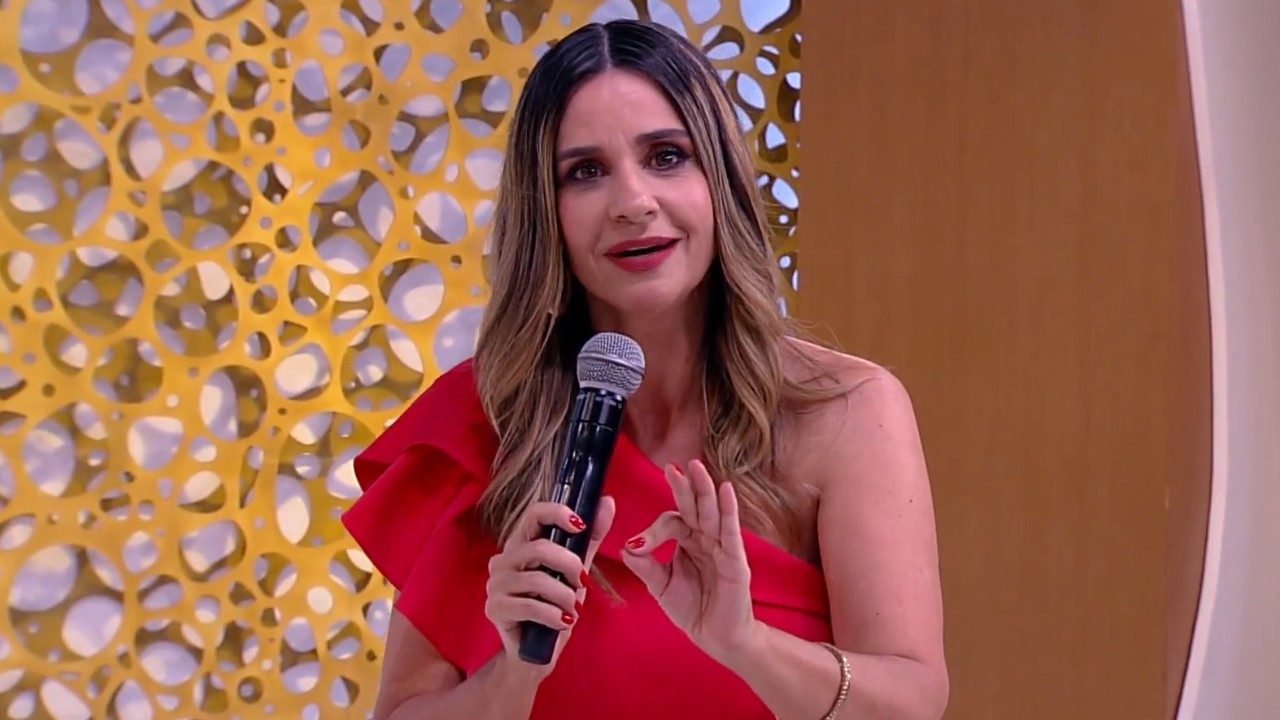 Repórter Simone Garuti no programa Superpop, usa vestido vermelho de um ombro só, segura microfone preto, fundo amarelo vazado