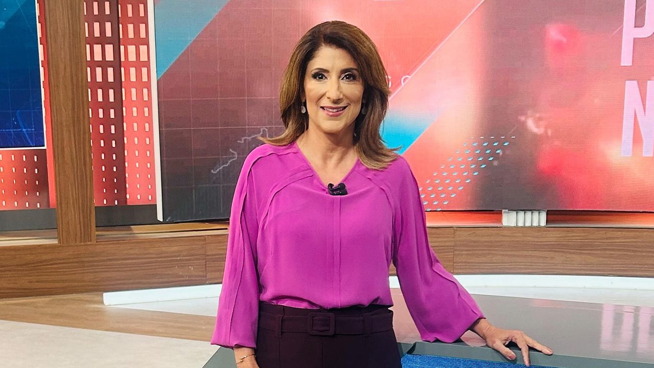 Simone Queiroz sorri ao lado da bancada no estúdio do SBT Brasil em frente ao telão do telejornal
