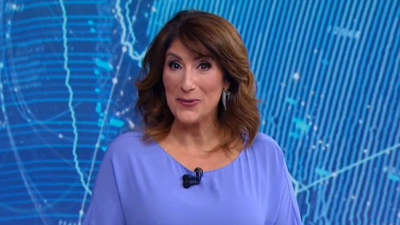 Simone Queiroz apresenta telejornal em estúdio com fundo azul de mapa digital e usa blusa lilás e microfone de lapela