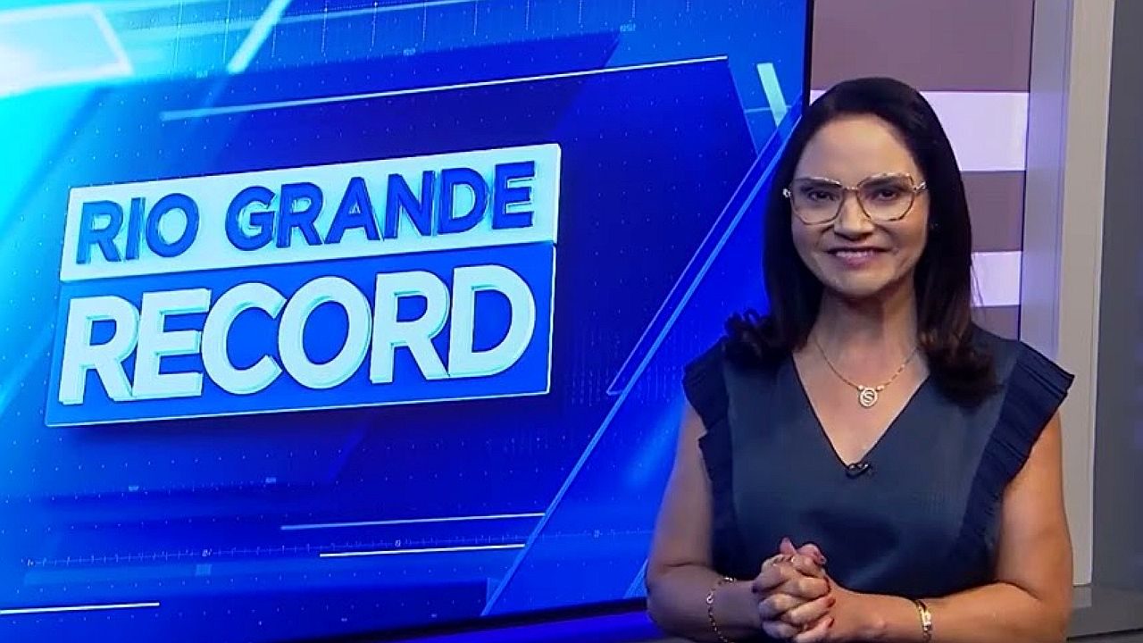 Uma mulher está em um estúdio de televisão, sorrindo enquanto apresenta um programa. Ela usa óculos, uma blusa escura sem mangas e um colar delicado. Suas mãos estão juntas à frente do corpo, em postura profissional. Ao lado dela, há um grande painel azul com o texto “RIO GRANDE RECORD”, indicando um telejornal ou programa regional. O cenário tem iluminação forte e elementos gráficos típicos de transmissão televisiva.
