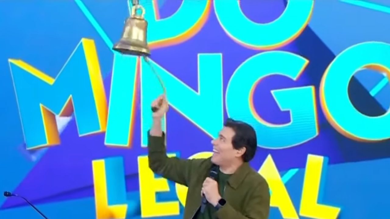 A imagem mostra Celso Portiolli sorrindo enquanto toca um sino dourado, segurando um microfone na outra mão. Ao fundo, há um grande painel colorido com as palavras Domingo Legal em letras grandes e vibrantes, mostrando que a cena ocorre no palco do famoso programa de televisão brasileiro. O clima é alegre e descontraído, característico das atrações de auditório