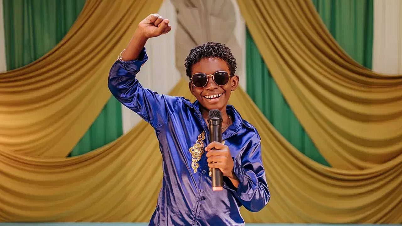 Menino de óculos escuros segura microfone e levanta o braço durante apresentação em palco colorido