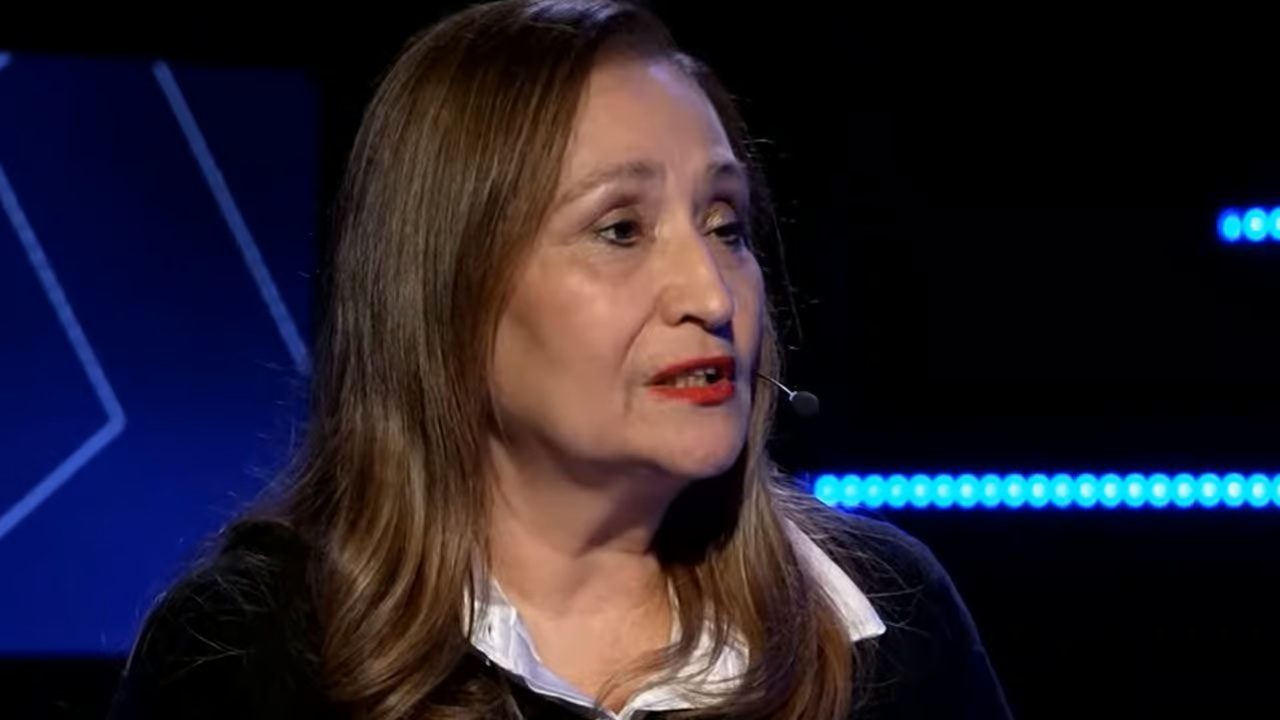 A imagem mostra uma mulher de cabelos castanhos e lisos, usando batom vermelho e roupa preta com gola branca. Ela está falando em um estúdio escuro, iluminado por luzes azuis de fundo, e usa um microfone de lapela próximo ao rosto, sugerindo que participa de uma entrevista ou programa de televisão