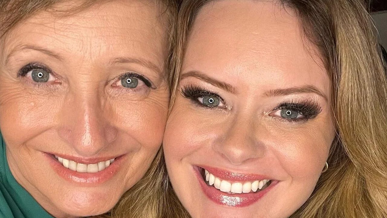 Close-up de duas mulheres sorrindo, com os rostos bem próximos, olhando diretamente para a câmera. À esquerda, uma mulher aparentando mais idade, com pele clara, olhos claros e rugas suaves ao redor dos olhos e do sorriso. À direita, uma mulher mais jovem, também de pele clara e olhos claros, com maquiagem marcante nos olhos e cílios longos. Ambas demonstram expressão alegre e afetuosa, sugerindo proximidade e carinho.