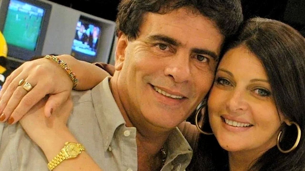 Homem e mulher sorriem abraçados enquanto posam para foto em ambiente interno, com televisores ao fundo sugerindo estúdio ou sala com monitores.
